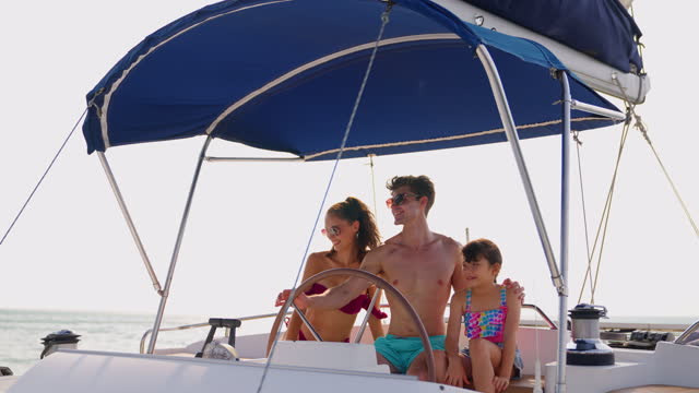 Caucasian happy family driving yacht outdoors on the sea during summer. Young beautiful couple in bikini hanging out and spending time with daughter sailing boat during holiday vacation trip together.