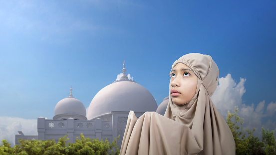 Asian Muslim girl in a hijab raising her hands and praying with a mosque and blue sky background