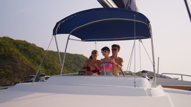 Caucasian happy family driving yacht outdoors on the sea during summer. Young beautiful couple in bikini hanging out and spending time with daughter sailing boat during holiday vacation trip together.