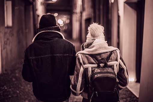 A couple of young people are captured walking hand in hand down a narrow alley lit up by streetlights at night.