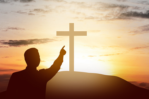 Silhouette of businessman pointing Christian cross on the hill with sunset sky background