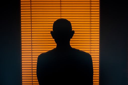 A man's shadow is projected onto a wall and window blinds at sunrise.