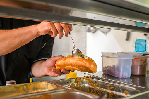 Close up shot of a professional male chef preparing a hot dog to go. He is using a long spoon to put the toppings.