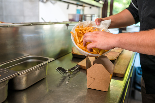 Male hands preparing fresh fries and placing them on a carton box to be taken away. Indoor shot at a food truck kitchen.