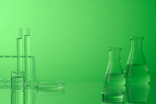 Laboratory glassware with liquid on green background. Test tubes, beaker, petri dish and erlenmeyer flask containing colorless liquid. Blank space to place your design