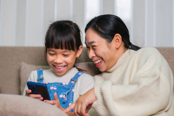 istock Young mother and her little daughter are using smartphone and smiling while sitting on sofa at home. Cheerful little girl using smartphone while relaxing with mom at home. Kids and Technology 1490837626