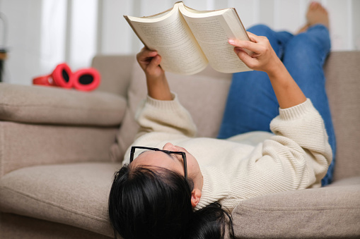 Happy Asian woman lying on the sofa and reading a book indoors. Lifestyle, free time and relaxation.
