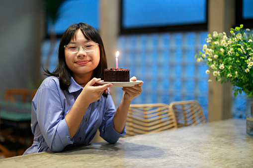 A cheerful Asian girl is holding her birthday cake with a lit candle, smiling warmly as she looks directly at the camera.