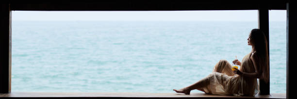 Barefoot young woman in silk flowing dress relaxing by coast with glass of fruit cocktail