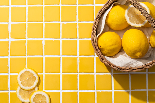Top view of bamboo basket with beige fabric containing a lot of fresh lemon. Some slices of lemon decorated on yellow tile background. Scene for advertising with lifestyle concept