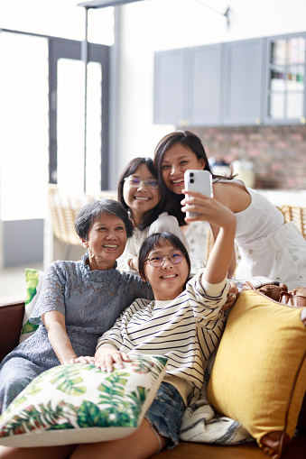 The granddaughter is using a smartphone to take a selfie with her grandmother, mother, and younger sister. They are all smiling happily, exemplifying the beautiful bond within a three-generation family.