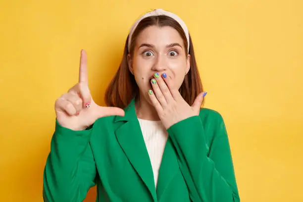 Shocked amazed woman wearing green jacket posing isolated over yellow background showing looser gesture looking at camera with surprised eyes covering mouth with palm.
