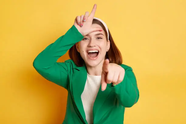 Funny laughing woman wearing green jacket posing isolated over yellow background showing looser gesture pointing at you expressing positive emotions.