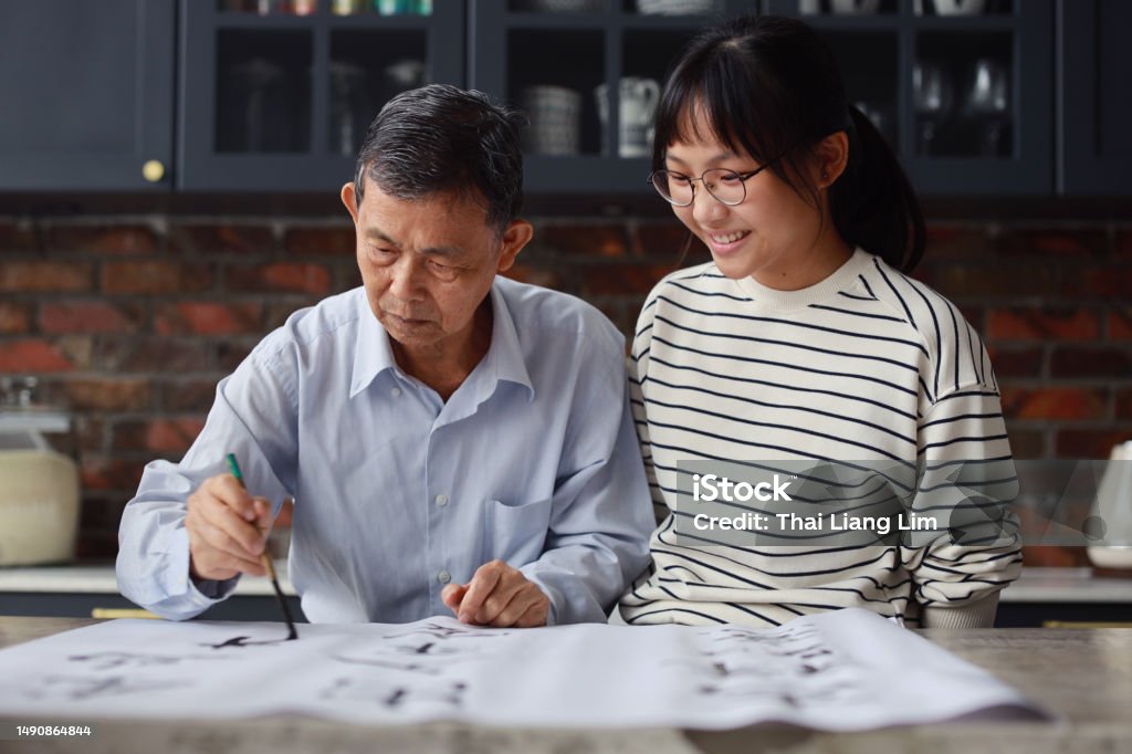 An Asian grandfather is seen teaching and showing his calligraphy skills to his granddaughter at home. The atmosphere is filled with warmth and love as the family members engage in this traditional Chinese activity together. This scene represents not only the passing down of cultural traditions but also the value of family and the importance of spending time together. It also promotes the concepts of youth education and learning, as well as an active and healthy lifestyle for seniors. Chinese Script Stock Photo