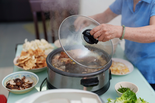 High angle view, a senior woman is seen diligently preparing a steamboat meal for her family at home. She carefully adds an assortment of vegetarian ingredients to the bubbling hot pot, ensuring a delicious and nutritious cooking experience.