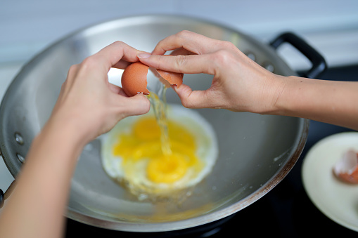 Cropped view of a woman's hands cracking an egg into a hot wok, preparing to fry it
