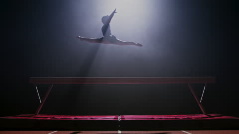 SLO MO LD Female balance beam gymnast doing a jump on the balance beam at night