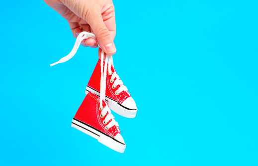 Close-up shot of a hand holding a pair of tiny red high-top canvas shoes by the laces against a bright blue background. Children's fashion or gift-related concept.