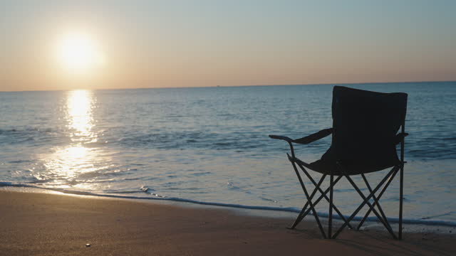 A folding Chair on the Seashore, facing the Sun. We meet the Dawn on the Sandy beach. Slow motion