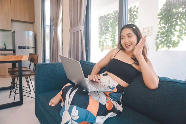 молодая азиатка проводит время дома, сидя на уютном диване в гостиной, проводя онлайн-конференцию на своем ноутбуке. - проводя поиск стоковые фото и изображения