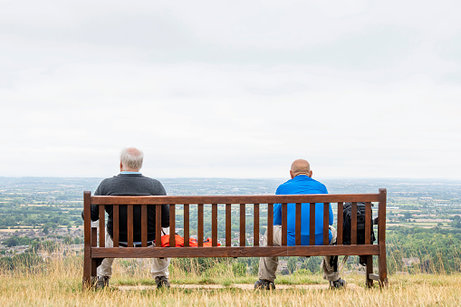 View from behind of two man in hiking gear and colorful clothing sitting on a bench on top of a hill with panoramic view over the Cotswolds landscape in the UK with an overcast sky