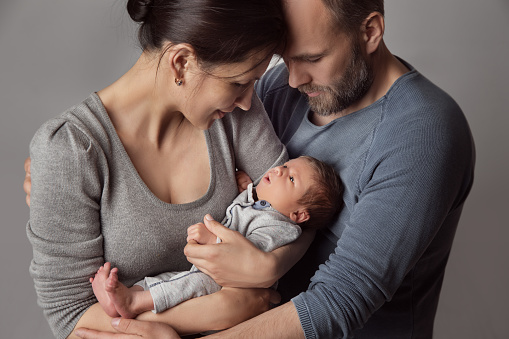 Parents holding Newborn Baby in Arms. Family Love. Father embracing Mother looking at infant Child over Gray Background. Children Care and Health