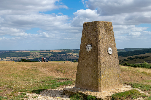 Low angle close up view of concrete pillar of the trig point on Cleeve Hill summit, Cheltenham, UK and marker for the public footpath Cotswolds Way with some golfers on the green in the background