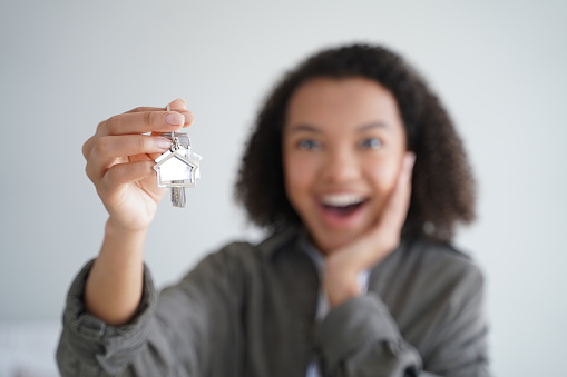 Excited woman showing keys of house, close-up focus on hand with key. Happy young girl tenant delighted with purchasing own apartment or relocating into new home. Real estate rental service concept.