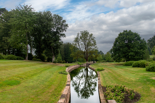 View over well-manicured landscape with stone walled river running through meadow and tree reflected in tranquility of the water along public footpath Cotswold Way near Chipping Sodbury, Bristol, UK