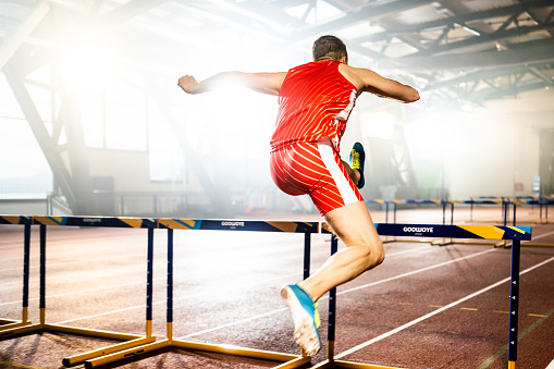Male caucasian athlete in sportswear running fast and jumping through hurdle while training at stadium track back view