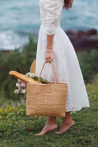 A stunning woman standing on a grassy lawn near the beach, wearing an ultra-chic vintage outfit with accessories and flowers in the beach bag. Stylishly embracing boho style for the perfect weekend look.