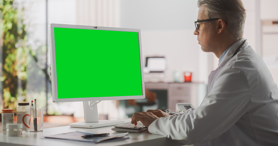 Healthcare Doctor Working on Desktop Computer with Green Screen Mock Up Display. Clinic Professional Using PC for Efficient Online Medical Work in Modern Office in Public Health Care Facility