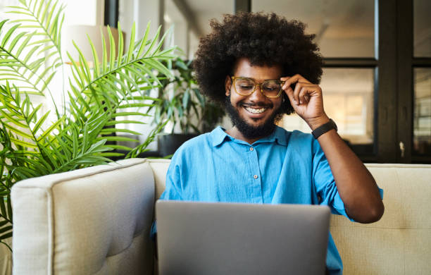 glücklicher, junger schwarzer mann mit einem afro, der auf der couch sitzt und auf den bildschirm seines laptops schaut. - shirt ethnic ethnicity one person stock-fotos und bilder