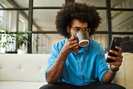 Young black man with a beard and an afro hairstyle sitting on a sofa scrolling through his phone whilst sipping on a hot beverage wearing casual clothing. Copy space, stock photo