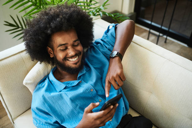 istock Young black man laying on a sofa scrolling on his mobile phone smiling and laughing whilst wearing a a blue shirt with copy space. Stock photo 1490904383