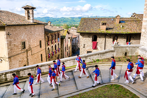 Gubbio, Umbria, Italy, May 15 -- A scene of the traditional and ancient Festa dei Ceri in the historic heart of the medieval town of Gubbio in Umbria. The celebration consists of a race of three wooden columns weighing almost 300 kg each, called Ceri, carried on the shoulders of dozens of bearers, on top of which are placed the statues of Sant'Ubaldo (Saint Ubaldo) protector of Gubbio, San Giorgio (Saint George) and Sant'Antonio Abate (Saint Anthony).\nThe race develops along the streets and alleys of Gubbio up to the summit of Monte Ingino where the Basilica of Sant'Ubaldo is located. This religious celebration, much loved by the whole community of Gubbio, is considered one of the oldest in Italy and in the world and its origins date back to the 12th century. In the photo: A group of young participants in the race and bearers of the Ceri walk through the stone alleys of Gubbio. Image in high definition quality.