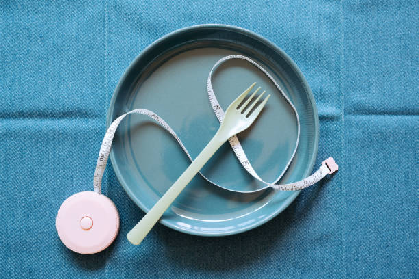 empty navy ceramic plate with fork and measuring tape on blue fabric table background. healthy food diet concept. top view - textile healthy eating instrument of measurement tape measure imagens e fotografias de stock