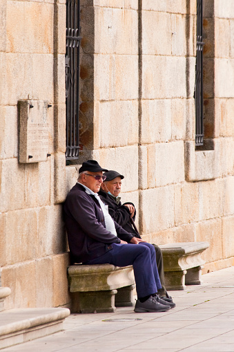 Cambados, Spain_ March 23, 2022: Senior men sitting and talking  on a stone bench in town square. Cambados town hall facade, Rías Baixas, Pontevedra province, Galicia, Spain.