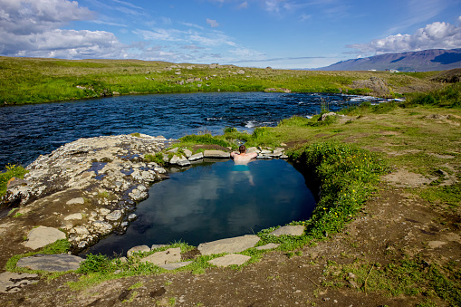 Man relaxing and bathing in natural geothermal heated hot pool, hot spring in Iceland in summer. Green rolling hills and blue sky on background. Hot spring is called Fosslaug hot pot.
