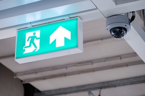 Fire exit sign with closed-circuit television (CCTV) camera installed on ceiling structure in the building. Security and safety system for public building