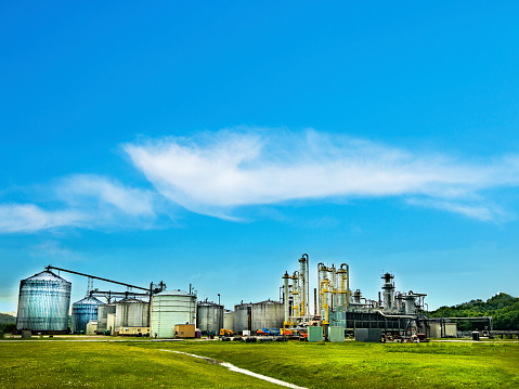 An Ethanol refinery factory is surrounded by a green field and blue sky. The image features silos and buildings surrounded by lush greenery.