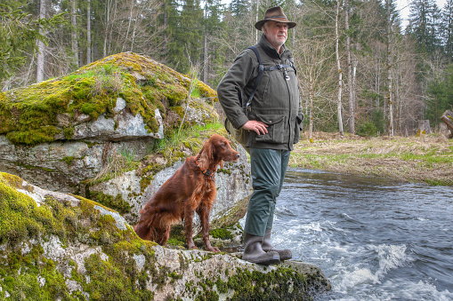 An elderly hiker and his trusty Irish Setter stand on a large rock jutting out of the water. Both carefully observe the surroundings and listen to the soothing sound of the river.