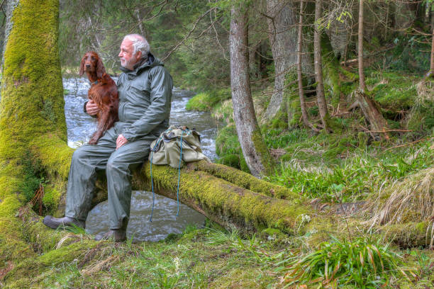 Serene Moments by the River.  A Hiker and his Irish Setter Companion Resting on a Riverside Log. stock photo