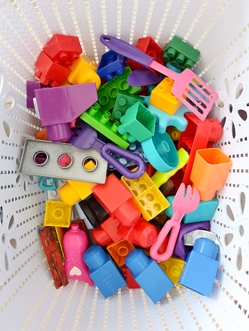 A white plastic basket full of colorful mixed toys to assemble and to play, in a kid's room.