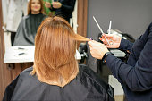 Red-haired woman sitting a front of the mirror and receiving haircut her red long hair by a female hairdresser in a hair salon, back view.
