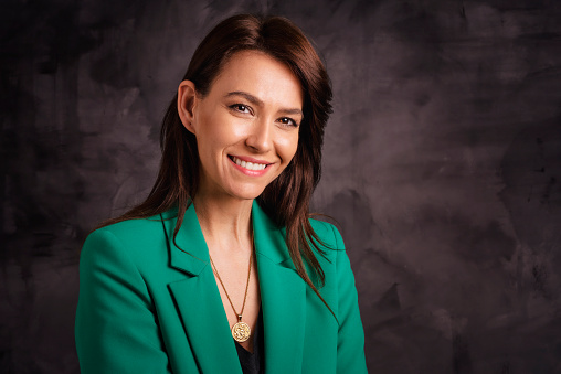 Headshot of an attractive middle aged woman cheerful smiling and looking at camera. Brunette haired female wearing blazer while sitting at isolated dark background. Copy space. Studio shot.