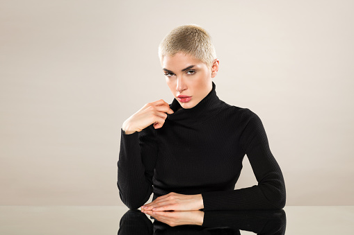 Portrait of serious young female in black turtleneck with short blond hair looking at camera against beige background