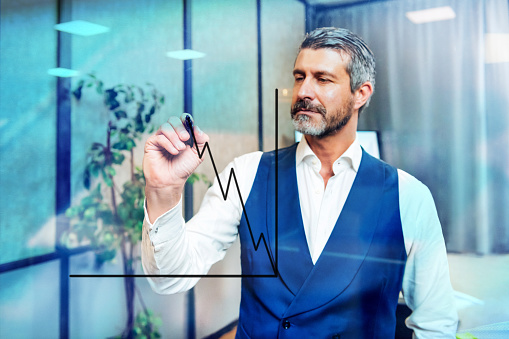 Through glass view of focused bearded male entrepreneur in white shirt and blue vest drawing chart on window with black marker