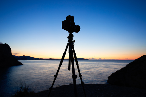 Photo camera silhouette on tripod at rocky beach with beautiful sunset in blue sea on seascape background.  Film analog photography