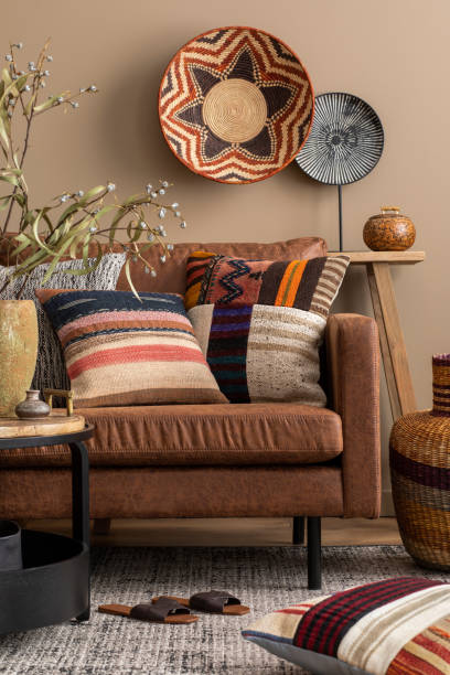istock Creative composition of living room interior with brown sofa, colorful pillows, patterned baskets, round coffee table, slippers, vase with eucalyptus and personal accessories. Home decor. Template. 1490957403